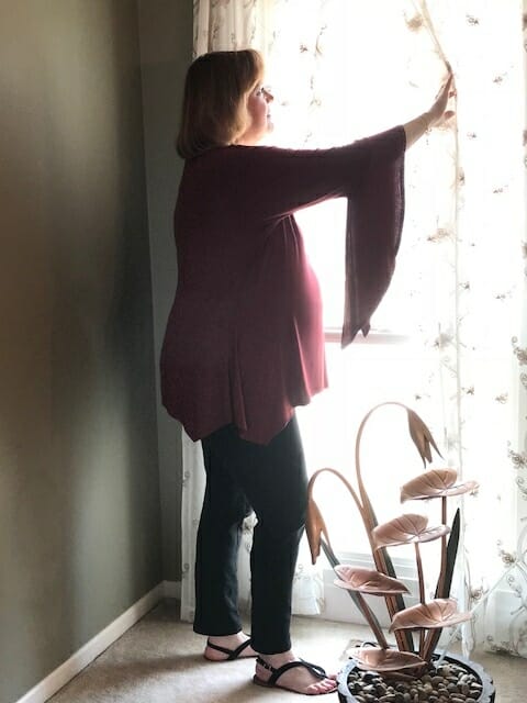 Side view of a Midnight Velvet customer by a sunny window, in a burgundy tunic with flowing sleeves, and black pant.