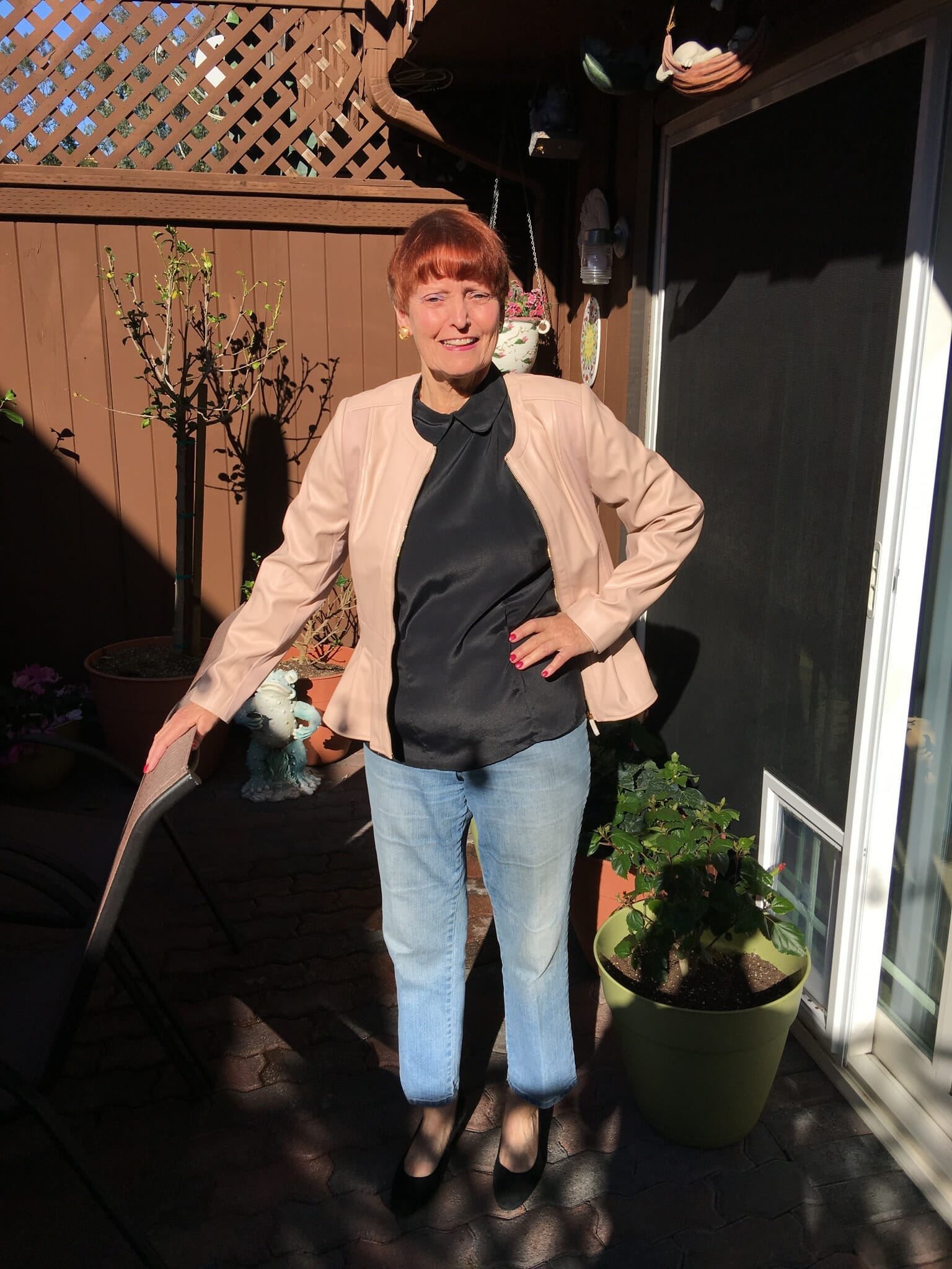 A smiling Midnight Velvet customer by a patio garden, in a blush peplum jacket, gray top, denim jeans and black pumps.