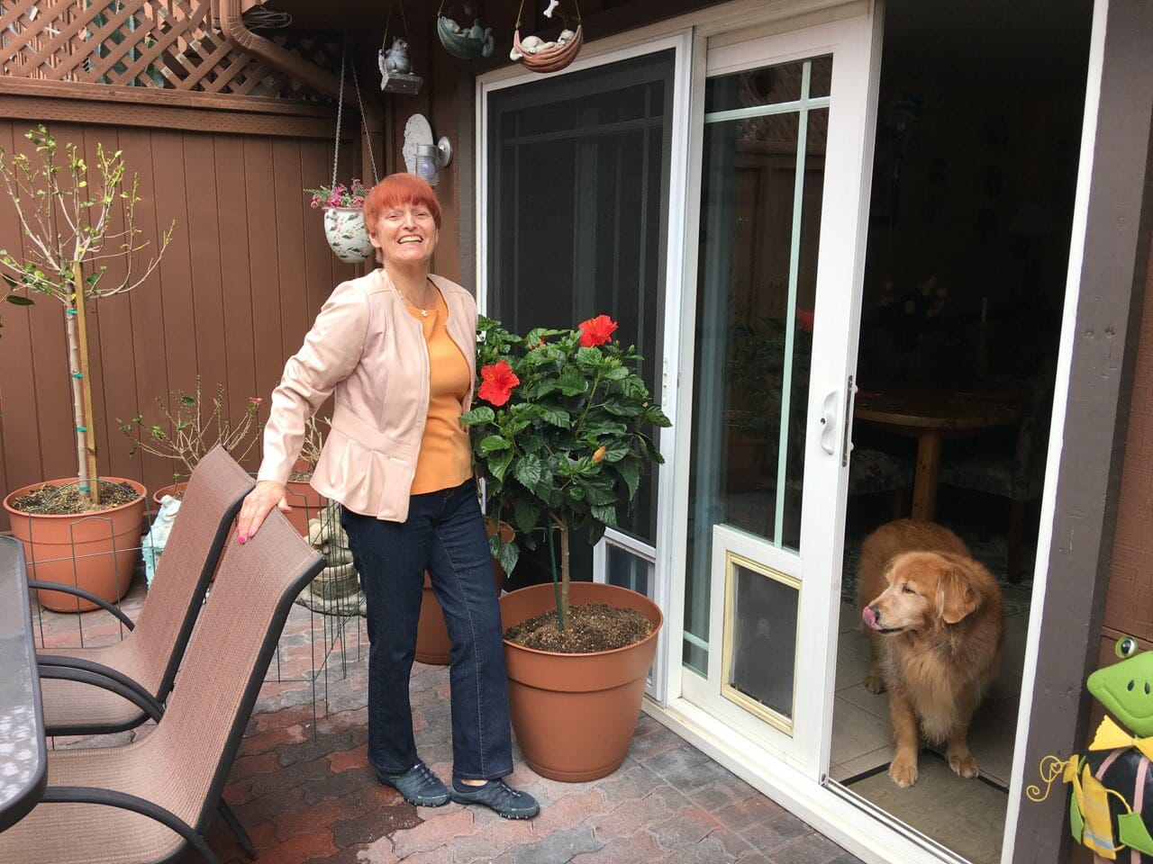 A Midnight Velvet customer and her dog by a patio garden, wearing a blush peplum jacket, peach top, and denim jeans.