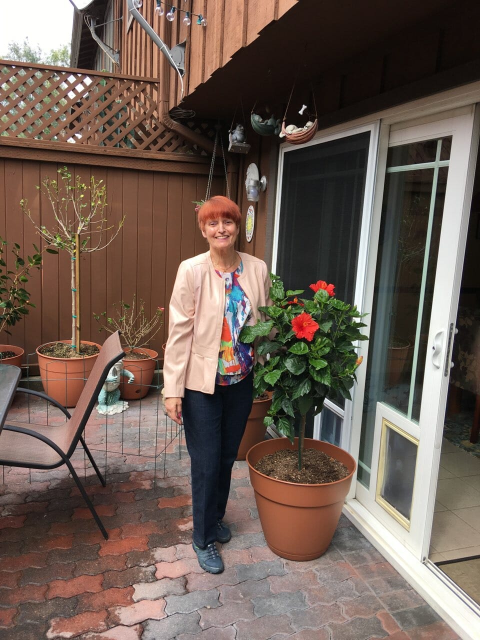 A smiling Midnight Velvet customer by a patio garden, wearing a blush peplum jacket ,colorful splash top, and denim jeans.