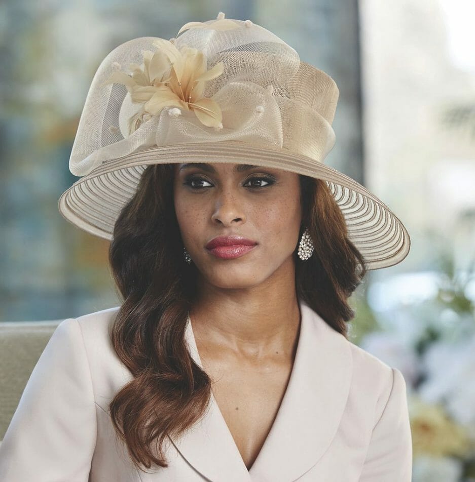 A brown eyed woman in a beige suit and fancy, wide brimmed hat with beige organza loops and beige lilies.