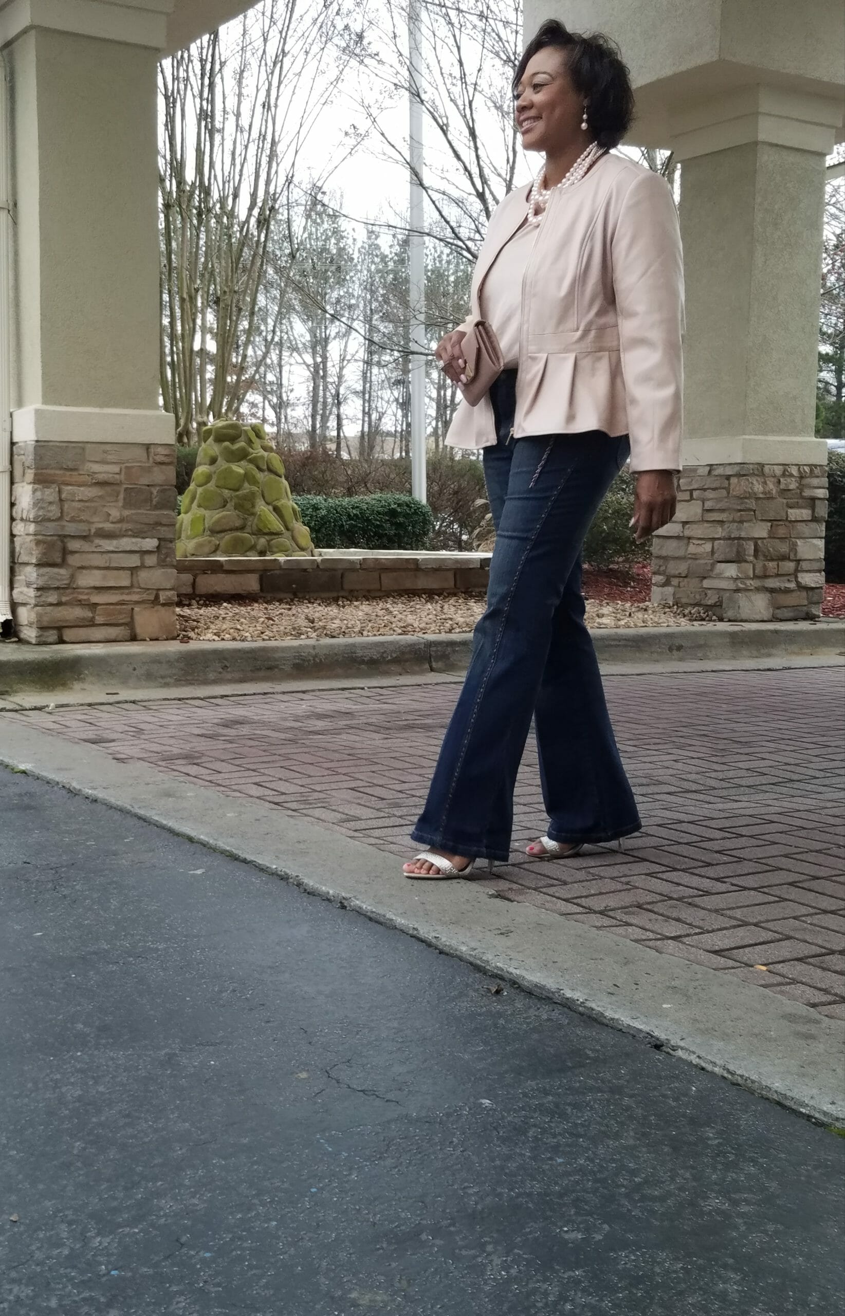 A Midnight Velvet customer outside in a blush top and peplum jacket set, jeans, sandals, and a pearl necklace.