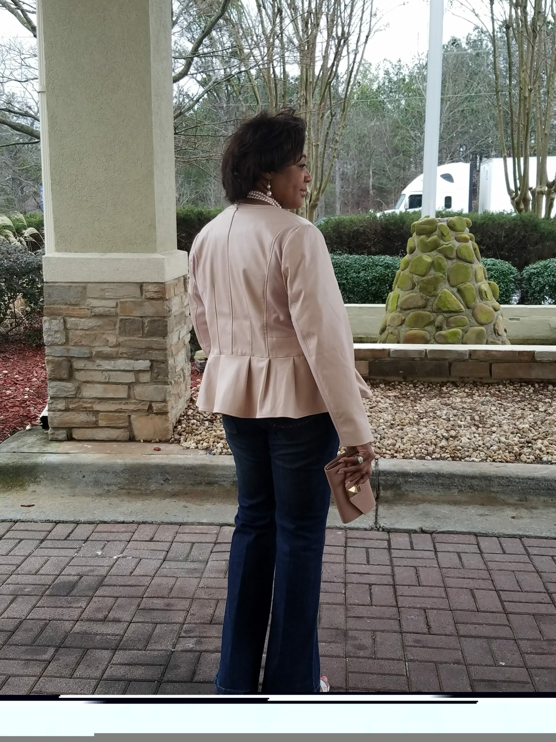 Back view of a Midnight Velvet customer outside in a blush peplum jacket, jeans, sandals, and a pearl necklace.
