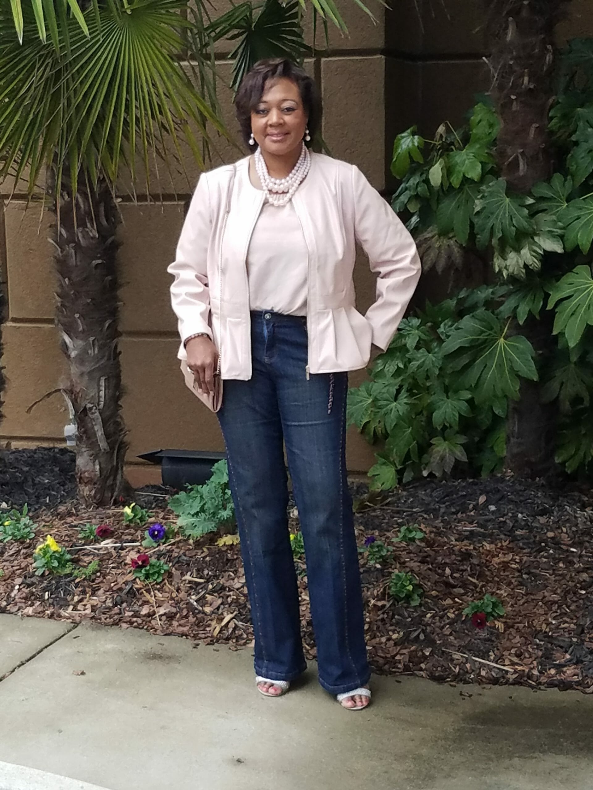 A Midnight Velvet customer by a palm tree, in a blush top and peplum jacket set, jeans, and a pearl necklace.