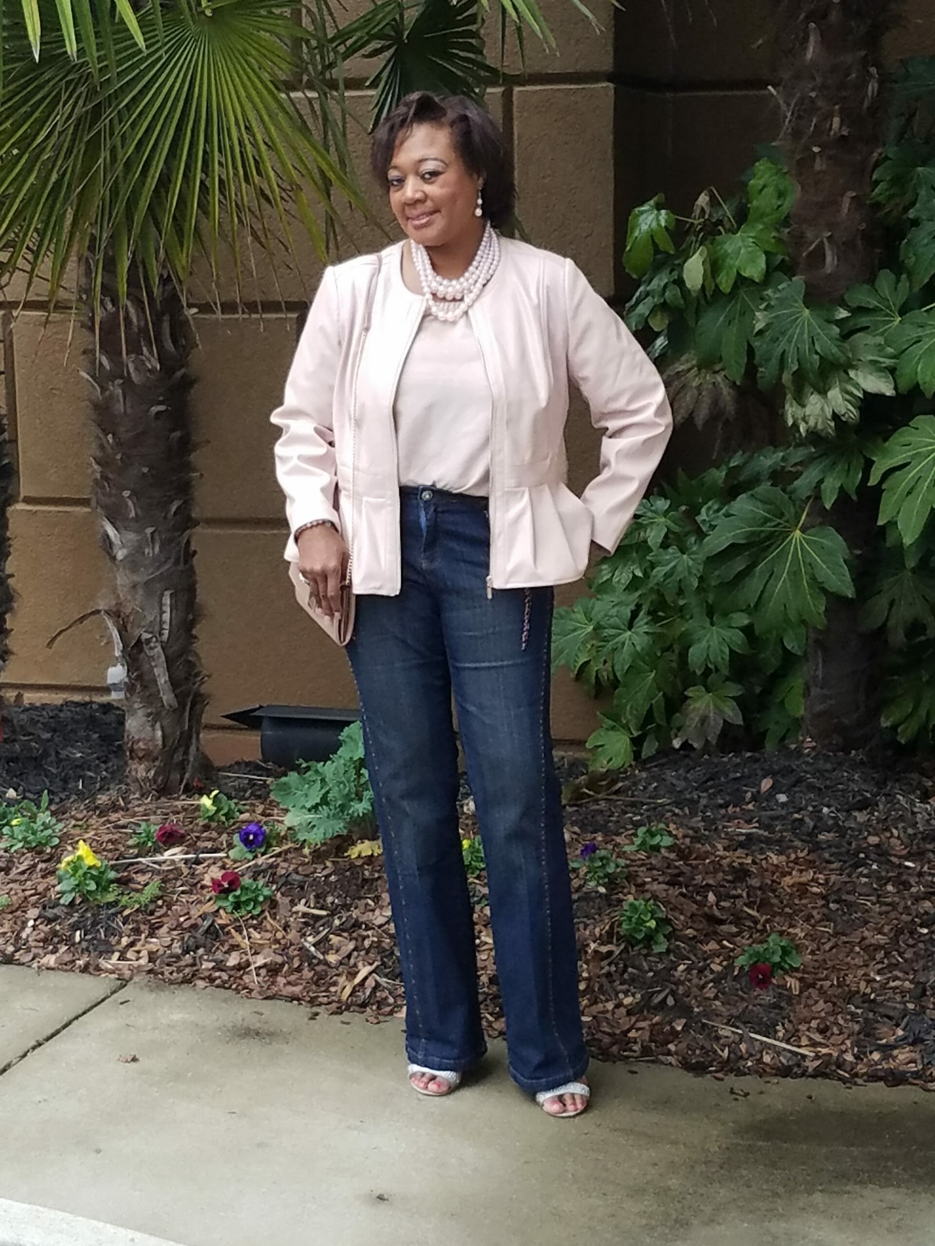 A Midnight Velvet customer by a palm tree, in a blush top and peplum jacket set, jeans, and a pearl necklace.