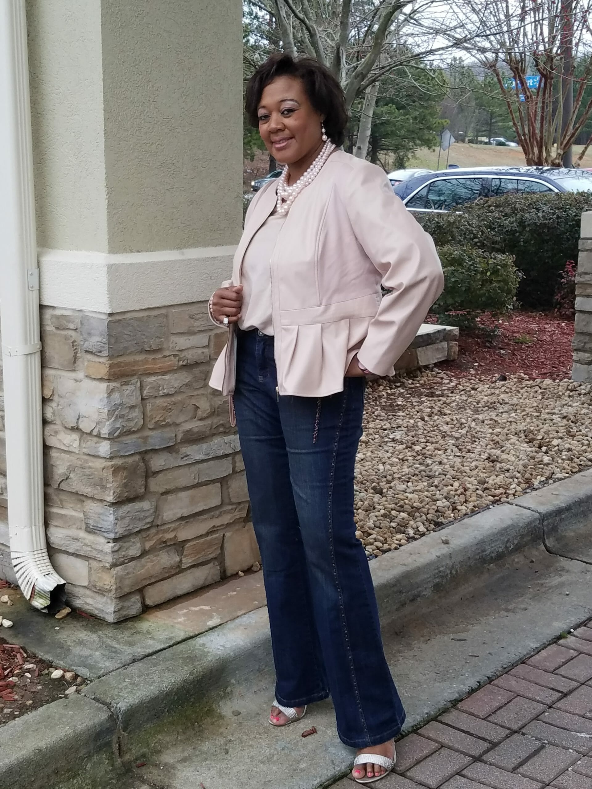 A Midnight Velvet customer by a rock column, in a blush top and peplum jacket set, jeans, sandals, and a pearl necklace.