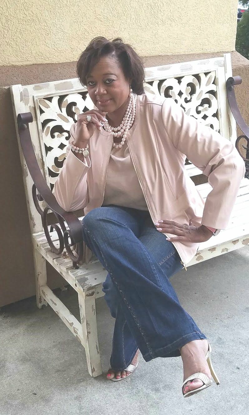 A Midnight Velvet customer seated on a bench, in a blush top and peplum jacket set, jeans, sandals, and a pearl necklace.
