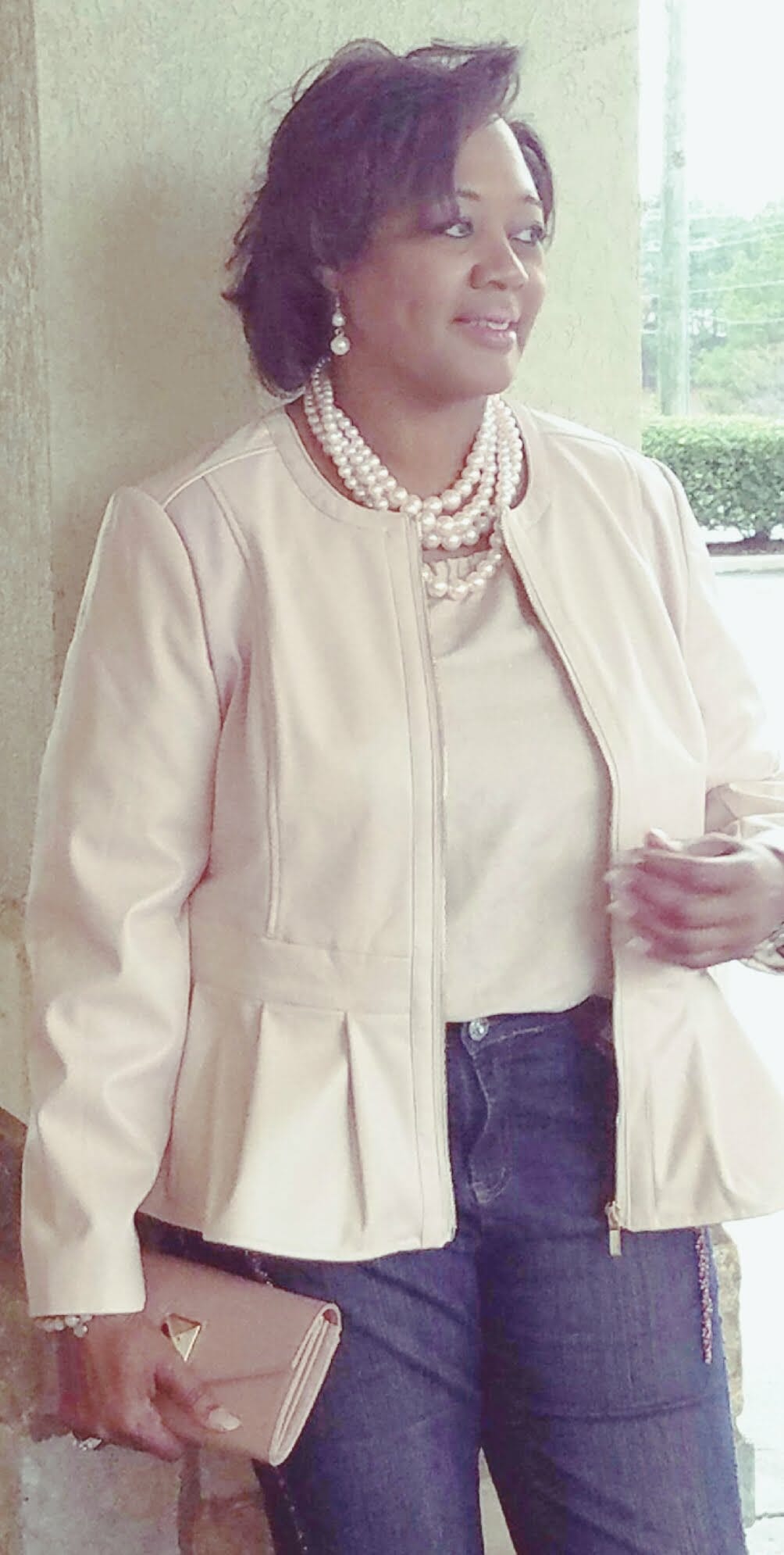 A Midnight Velvet customer in a blush top and peplum jacket set, jeans, a pearl necklace, and holding a blush clutch.