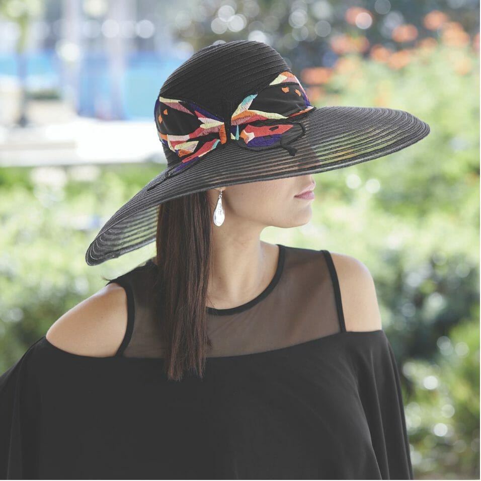 A woman in a black cold-shoulder top and a black woven hat with a wide brim and a multicolor fabric band and bow.