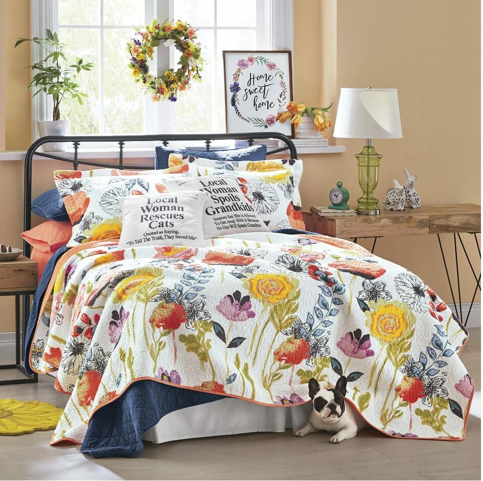 A bedroom with a colorful floral quilt, layered bedding, toss pillows with messages, a floral wreath, and a peeking pug dog.