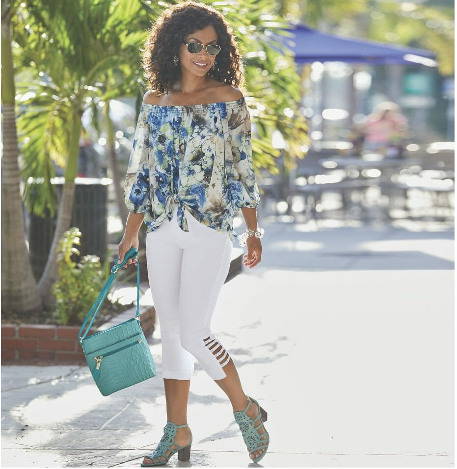 A black woman in sunglasses, a blue floral off-the-shoulder top, white capris, turquoise sandals, with a matching bag.
