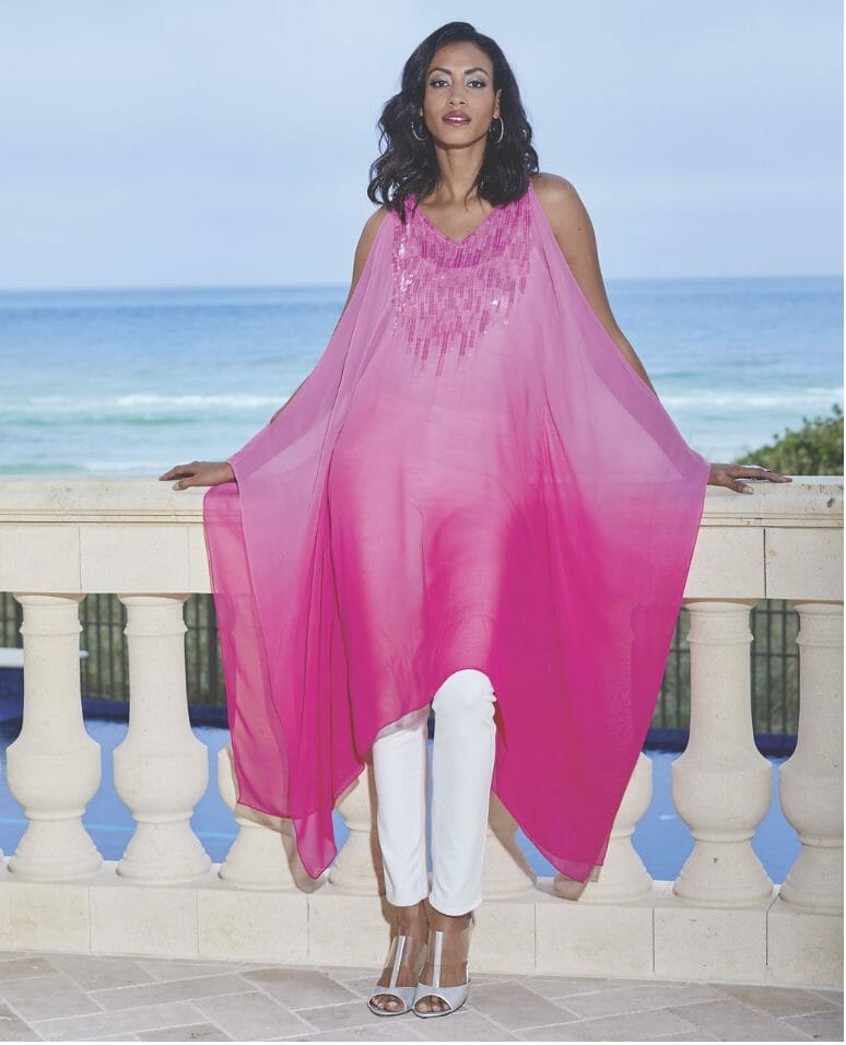 A woman on a seaside balcony, in a long, sheer magenta caftan with neckline sequins, and white skinny jeans and sandals.