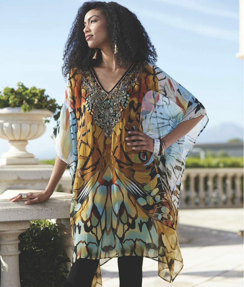 A black woman with long curly hair in a sheer Afrocentric caftan top with beaded neckline, and black leggings. 