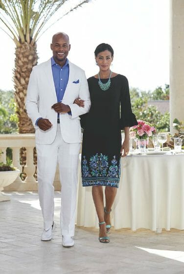 A balcony table setting with a man in a white suit, by a woman wearing a black and blue dress, and a turquoise necklace.