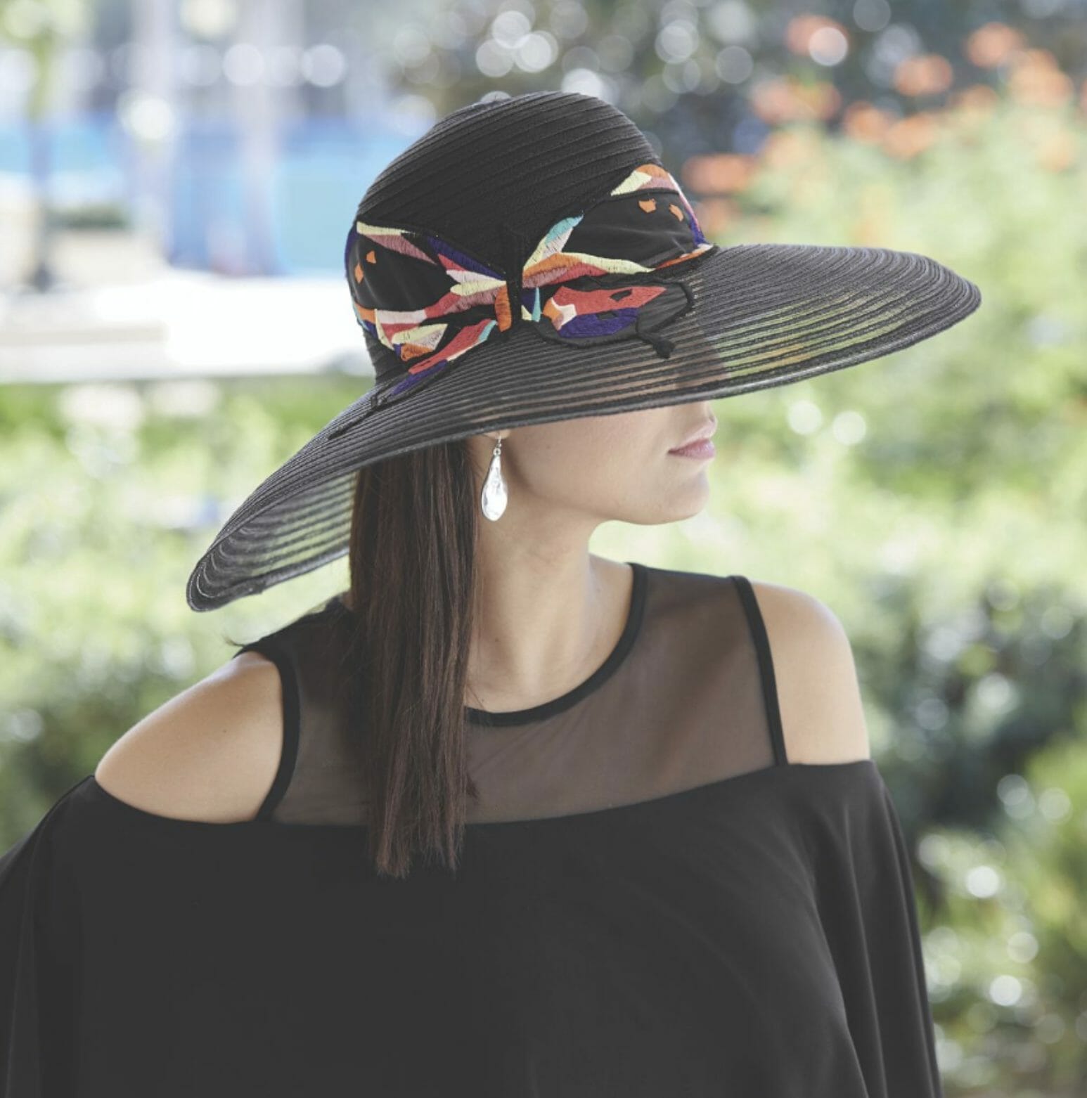 A woman in a black cold-shoulder top and a black woven hat with a wide brim and a multicolor fabric band and bow.
