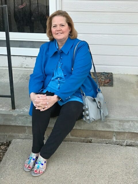 A Midnight Velvet customer seated on a step, in a blue shirt and jacket, black pant, turquoise sandals, with a gray bag.