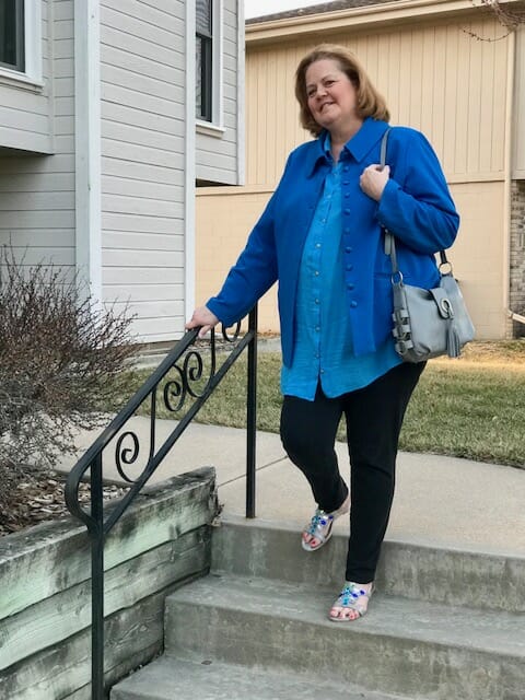 A Midnight Velvet customer going down steps, in a blue shirt and jacket, black pant, turquoise sandals, with a gray bag.