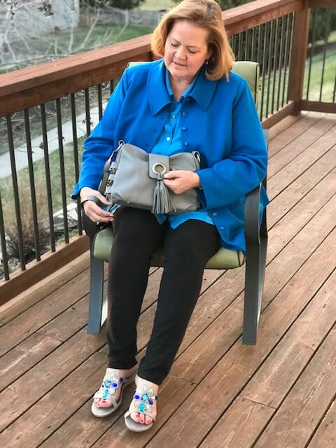 A Midnight Velvet customer seated on a deck, in a blue shirt and jacket, black pant, turquoise sandals, with a gray bag.