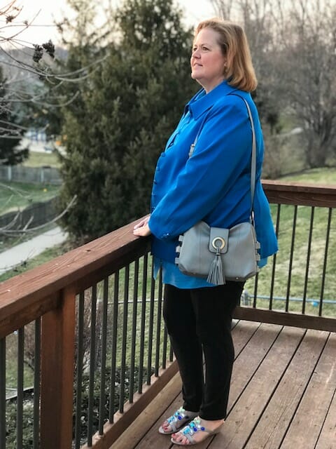 A Midnight Velvet customer standing on a deck, in a blue shirt and jacket, black pant, turquoise sandals, with a gray bag.