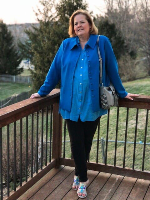 A Midnight Velvet customer standing on a deck, in a blue shirt and jacket, black pant, turquoise sandals, with a gray bag.