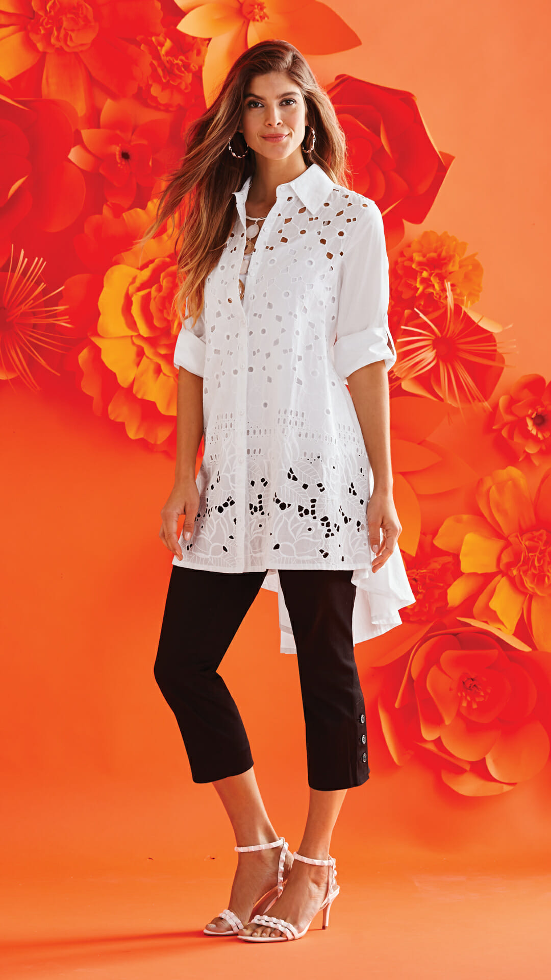 A woman in a white cutwork button-shirt tunic, black button capris, and white sandals, with an orange floral background.