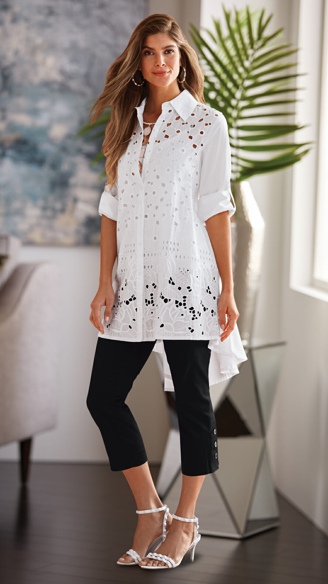A long-haired woman in a white cutwork button-shirt tunic with an asymmetrical hem, black capris, and white sandals.
