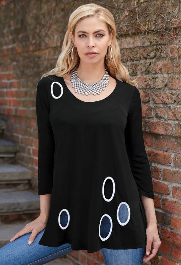 A blonde woman wearing a black scoop neck top with cutout circles, jeans, and a silver beaded collar necklace.