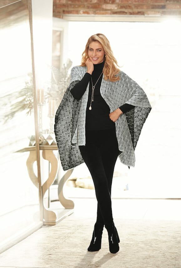 A smiling blonde woman in black leggings, booties and turtleneck, a silver woven blanket shawl, and a silver chain necklace.