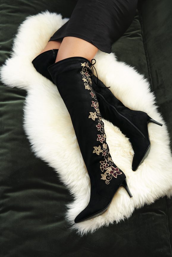 Lower portion of a woman in black over-the-knee boots with spike heels and brown and gold floral embroidery on the sides.