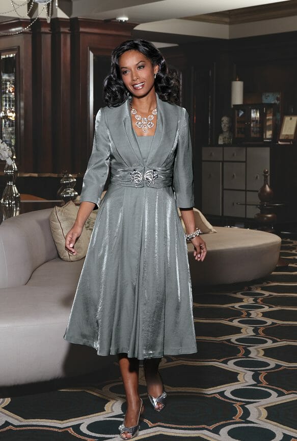 A smiling black woman in a silver jacket dress with a full skirt and jeweled clasp, silver sandals and a crystal necklace.