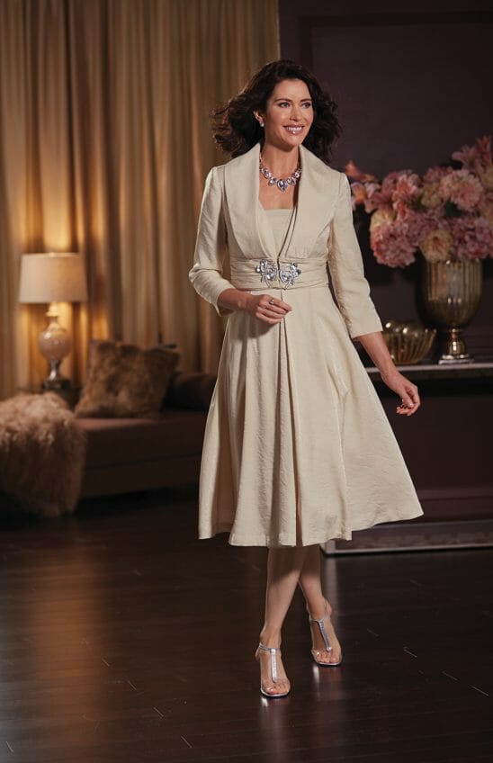 A smiling brunette woman in a beige jacket dress with a full skirt and jeweled clasp, silver sandals and a crystal necklace.