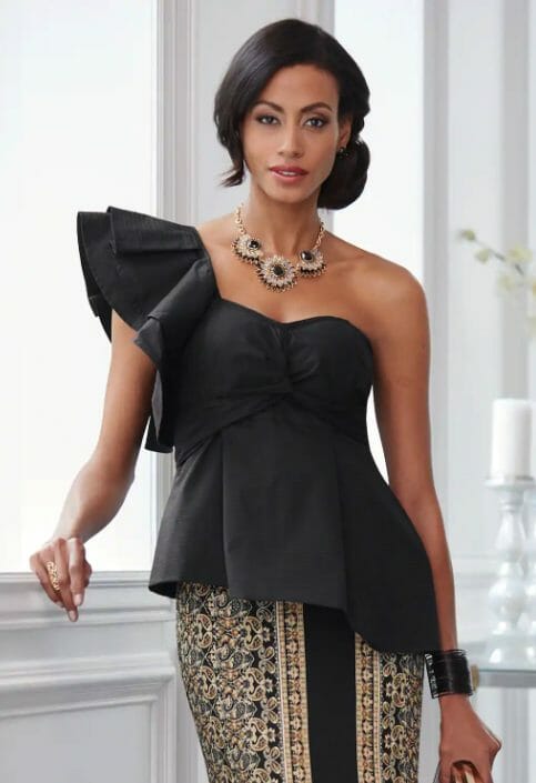 A woman in a fancy black top with a single ruffled sleeve, an asymmetrical hem, a tan and black baroque skirt and necklace.